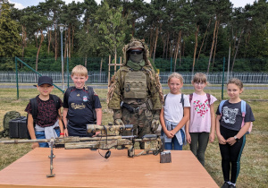 Żołnierz ubrany w strój maskujący pokazuje uczniom karabin snajperski.